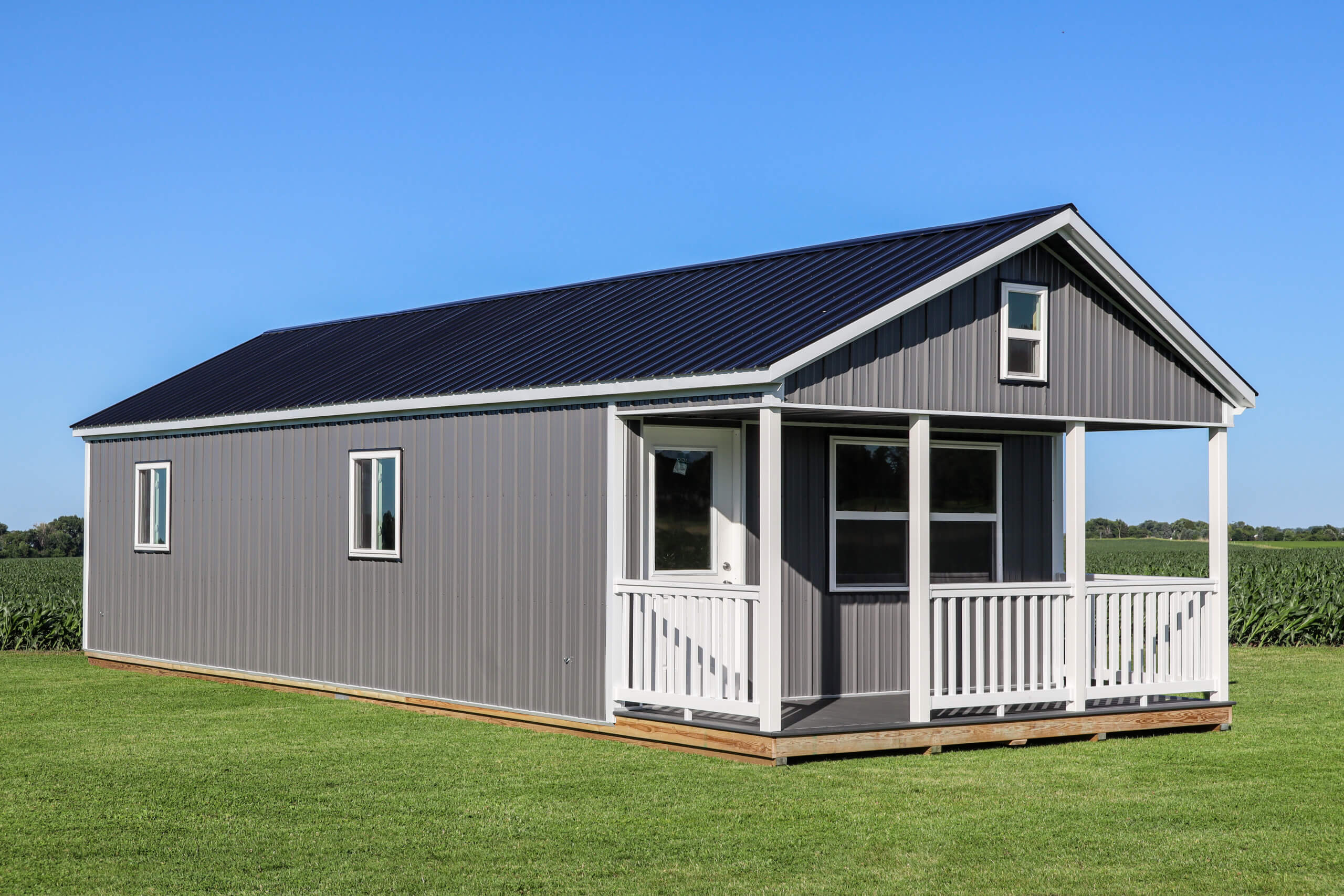 Gray tiny house with a black metal roof and white trim sits on a grassy lawn, featuring a small covered porch. Blue sky and fields are visible in the background.