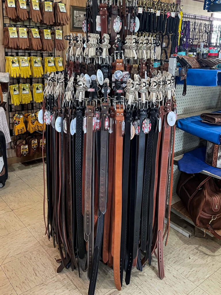 Penchem Tack Store Guthrie, KY A circular display rack holds various leather belts in black and brown; gloves and other accessories hang on the wall behind.