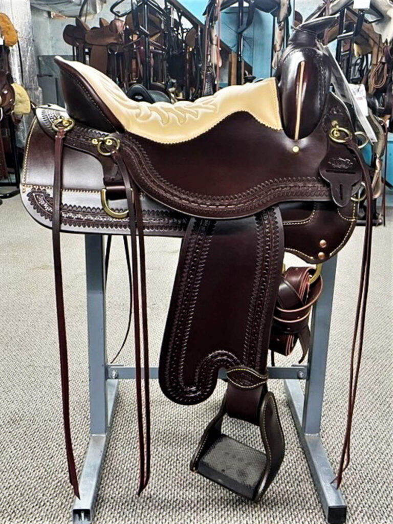 Penchem Tack Store Guthrie, KY A dark brown leather horse saddle with detailed stitching sits on a metal stand in a tack room, surrounded by other saddles and equipment.