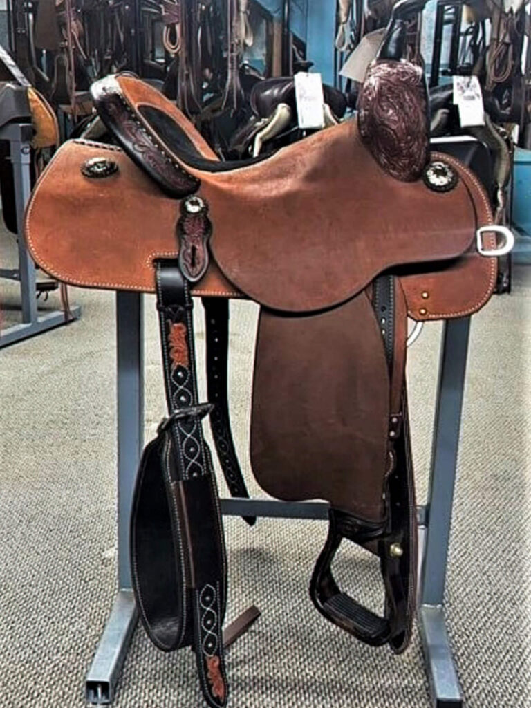 Penchem Tack Store Guthrie, KY A brown leather western saddle with intricate tooling is displayed on a metal stand in a store setting with other saddles in the background.
