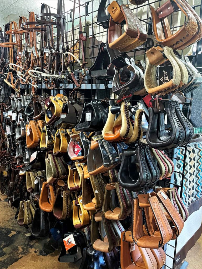 Penchem Tack Store Guthrie, KY A display of various saddlery items, including multiple types and colors of stirrups and leather straps, hanging on a metal grid wall in a tack shop.