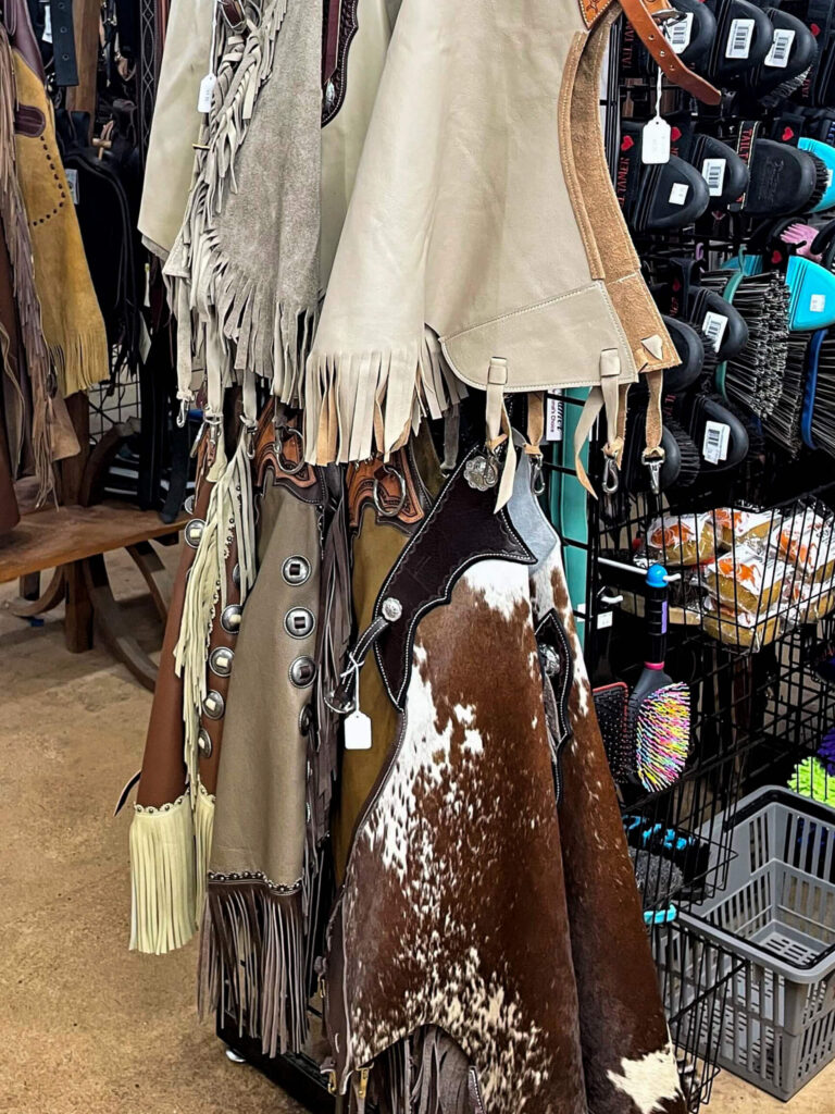 Penchem Tack Store Guthrie, KY Assorted leather and cowhide chaps with fringe and decorative buttons hang on a rack in a store, alongside various other equestrian gear.