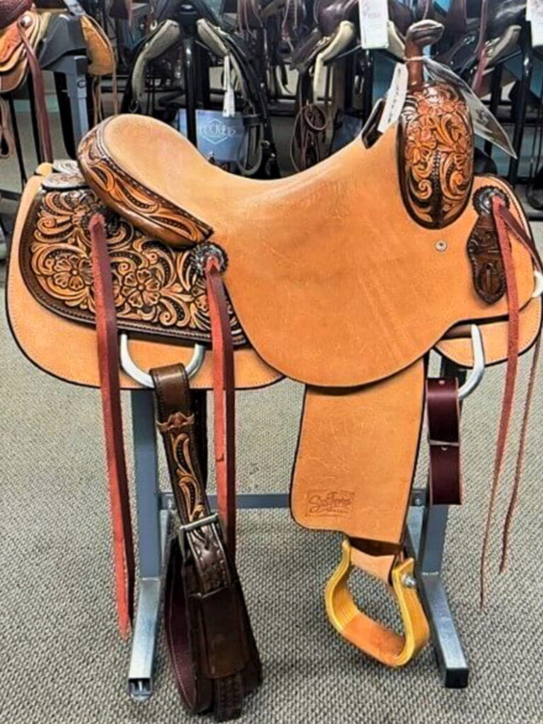 Penchem Tack Store Guthrie, KY A tan tooled leather western saddle with detailed floral patterns and yellow stirrups on a metal stand, displayed indoors among other saddles.