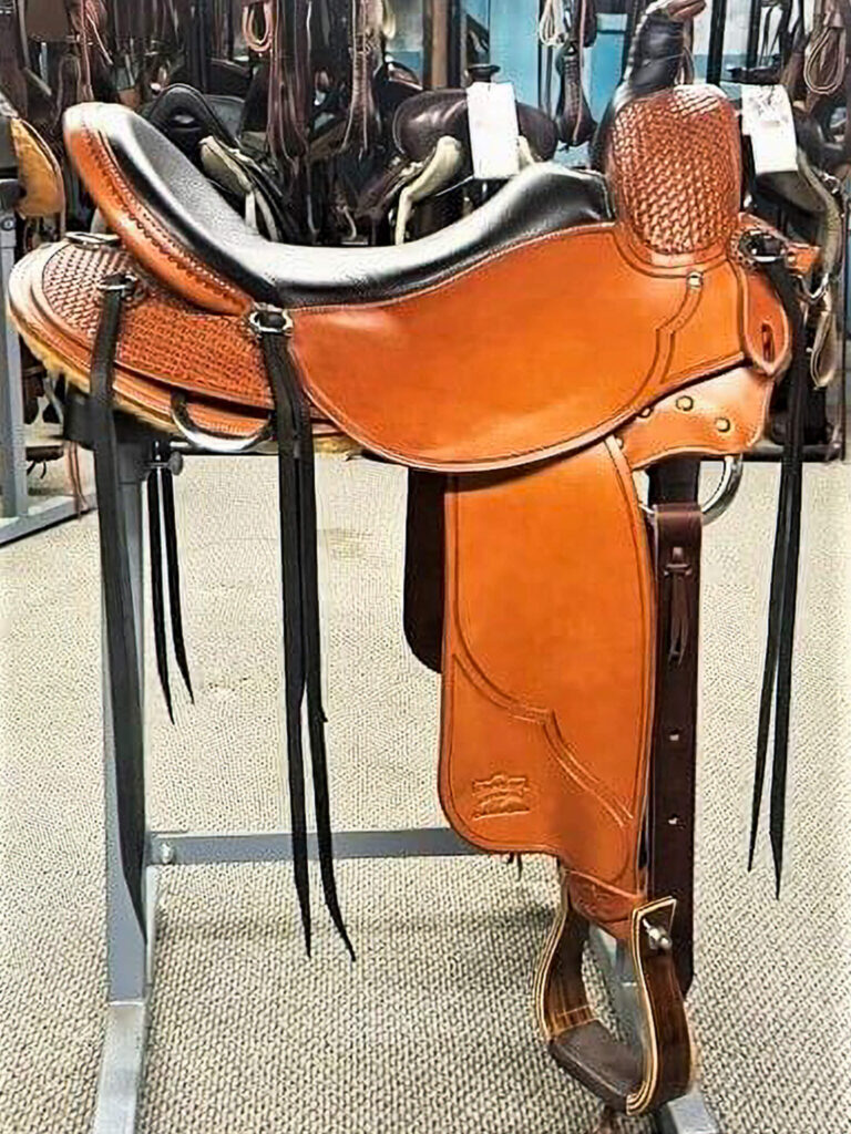 Penchem Tack Store Guthrie, KY A brown leather Western saddle with black accents is displayed on a metal stand inside a tack shop with other saddles in the background.