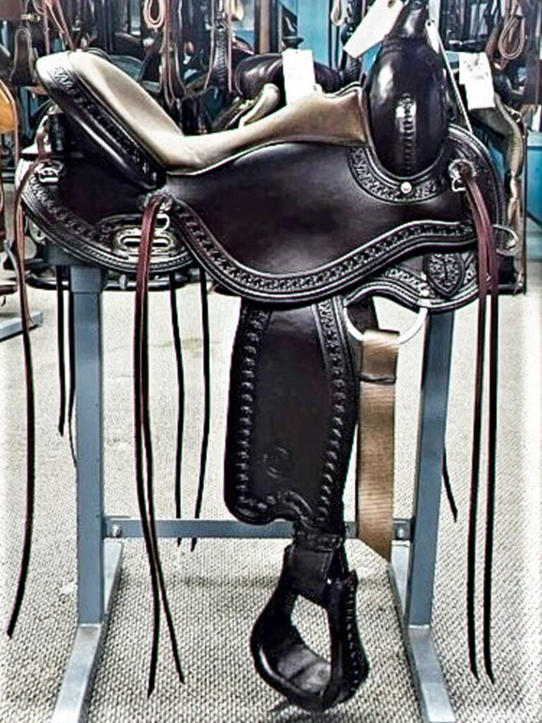 Penchem Tack Store Guthrie, KY A dark leather western saddle with decorative stitching sits on a metal stand in a tack room, surrounded by other horse gear.