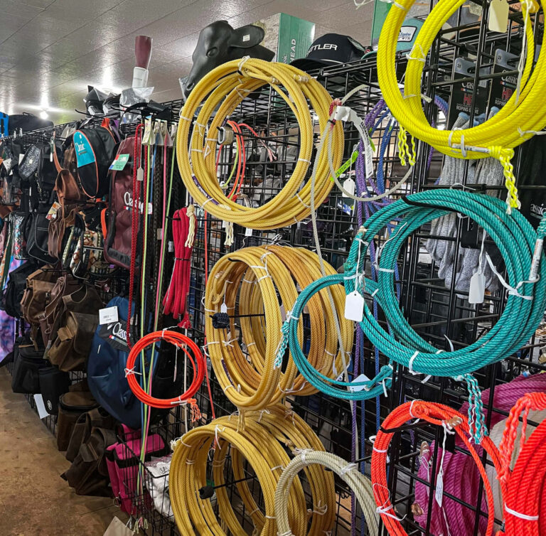 Penchem Tack Store Guthrie, KY Coiled ropes and lassos of various colors hang on a metal grid display inside a tack and supply store, with saddles and gear in the background.