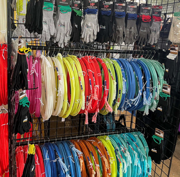 Penchem Tack Store Guthrie, KY Display of gardening gloves in various sizes and colors on the top rows, with coiled water hoses in multiple bright colors hanging below on a metal rack.