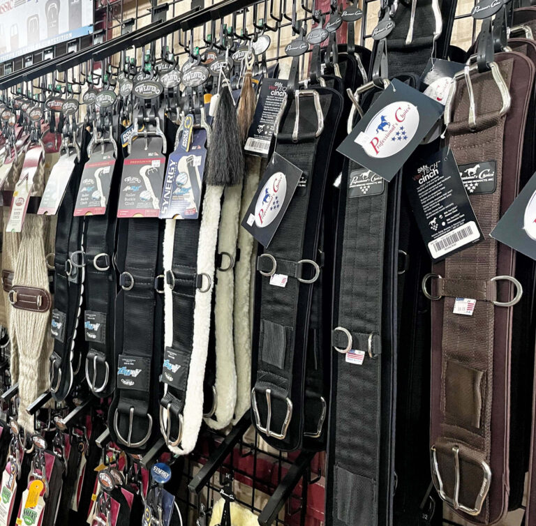 Penchem Tack Store Guthrie, KY A display of horse girths and cinches in various colors and materials hanging on hooks in a store aisle.