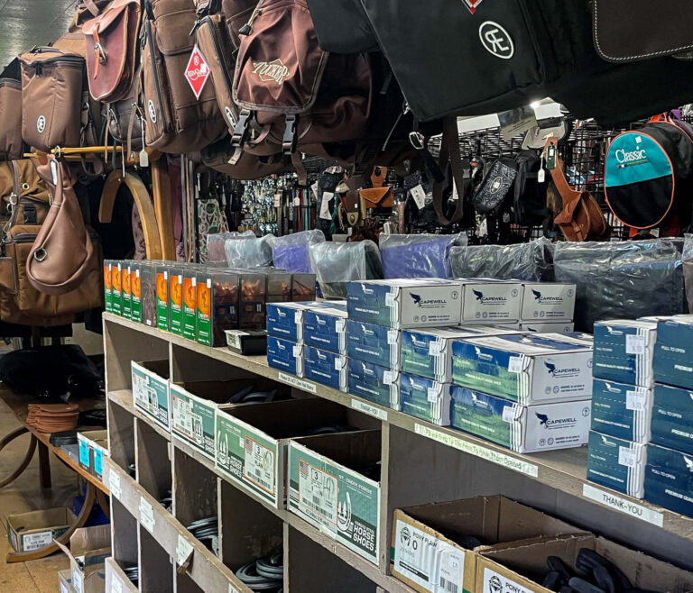 Penchem Tack Store Guthrie, KY Shelves stocked with various boxes and horse equipment, with saddlebags and riding gear hanging above in a tack shop.