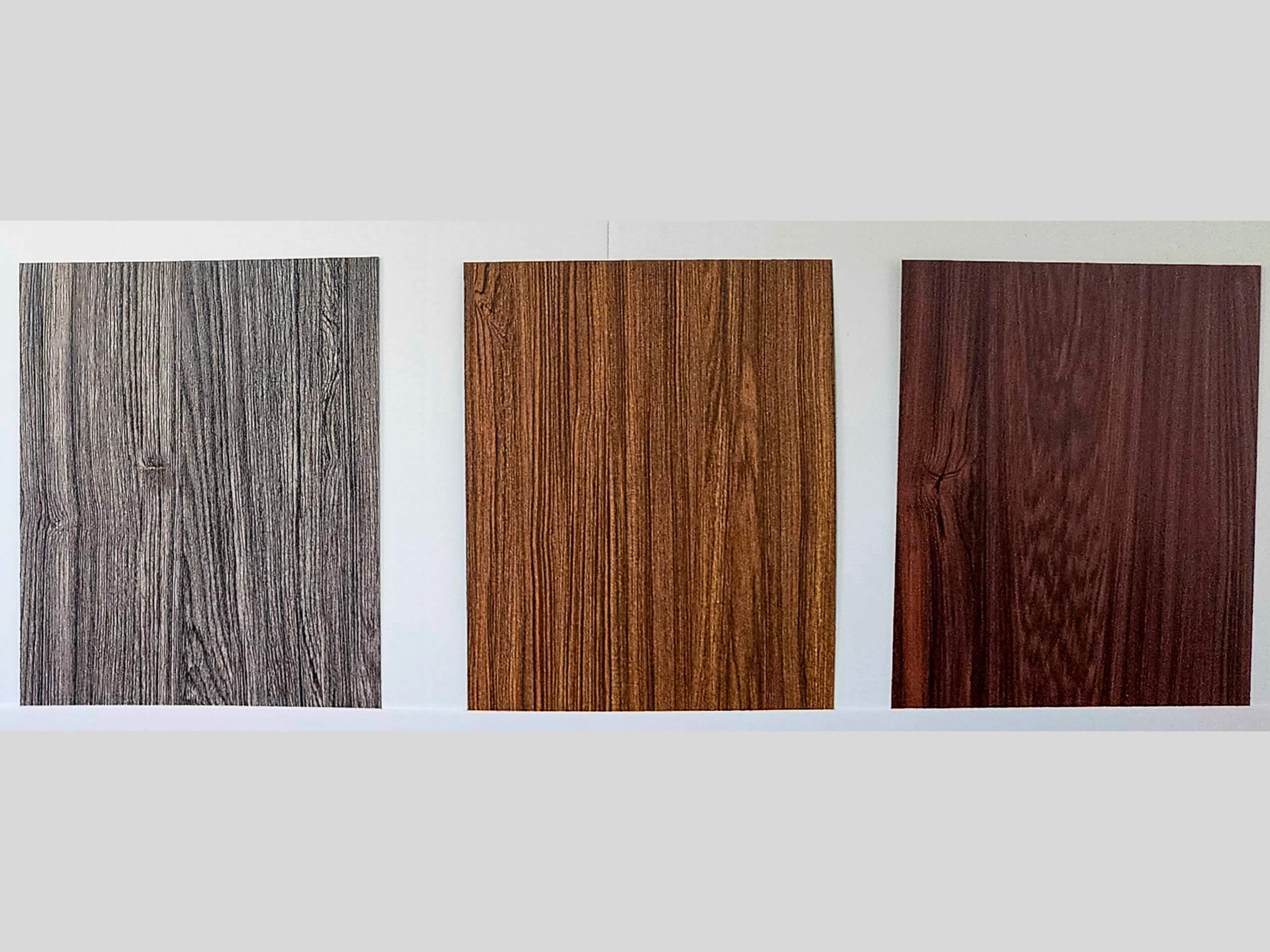 Sunset Metal Roofing and Siding Show Room Ebensburg, PA Three rectangular wood panels in gray, light brown, and dark reddish-brown tones are displayed vertically side by side against a plain light background.