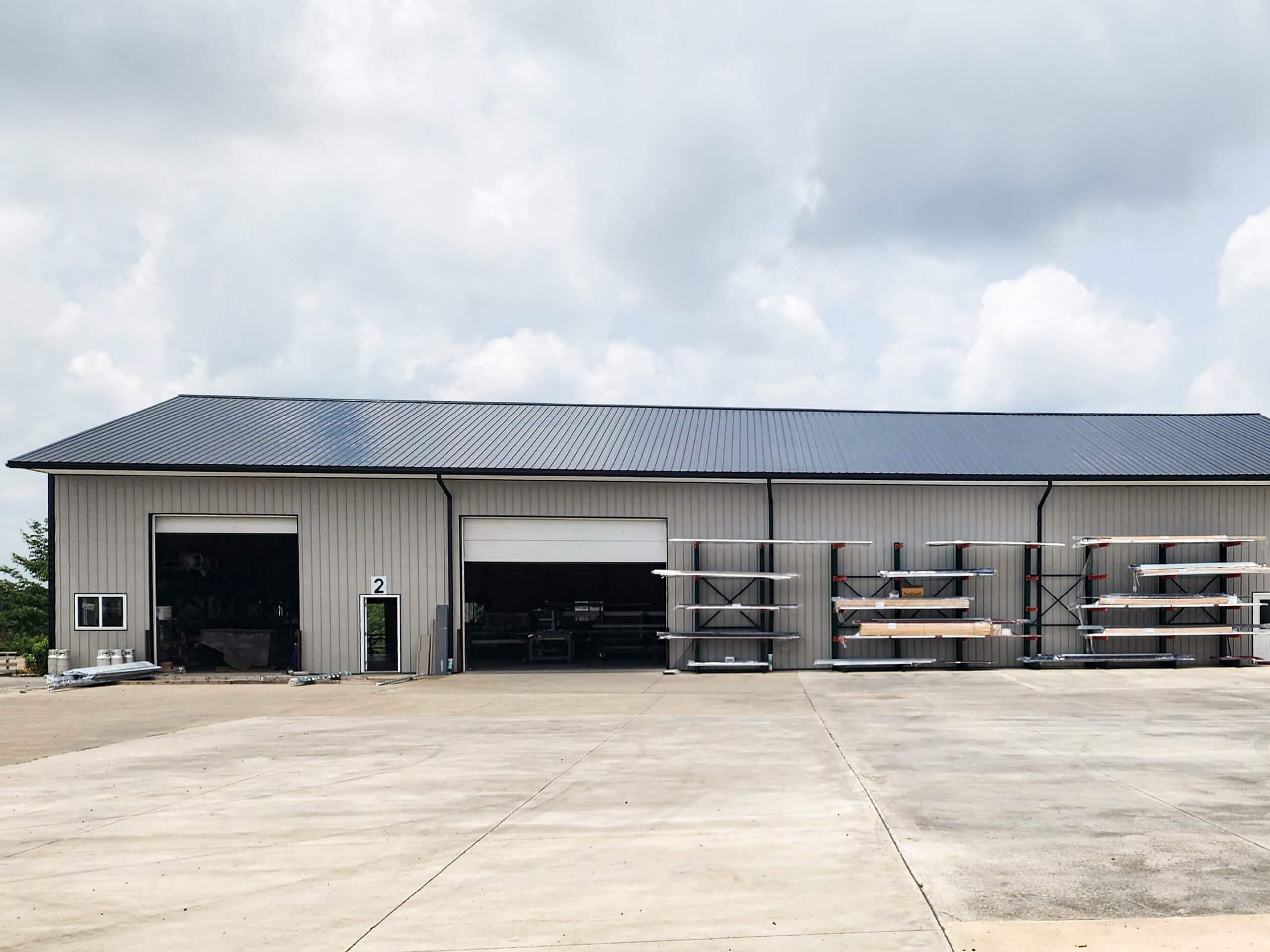 Sunset Metal Roofing and Siding Warehouse Ebensburg, PA Large metal warehouse with three open garage doors and shelves holding long metal beams outside on a concrete lot, under a cloudy sky.
