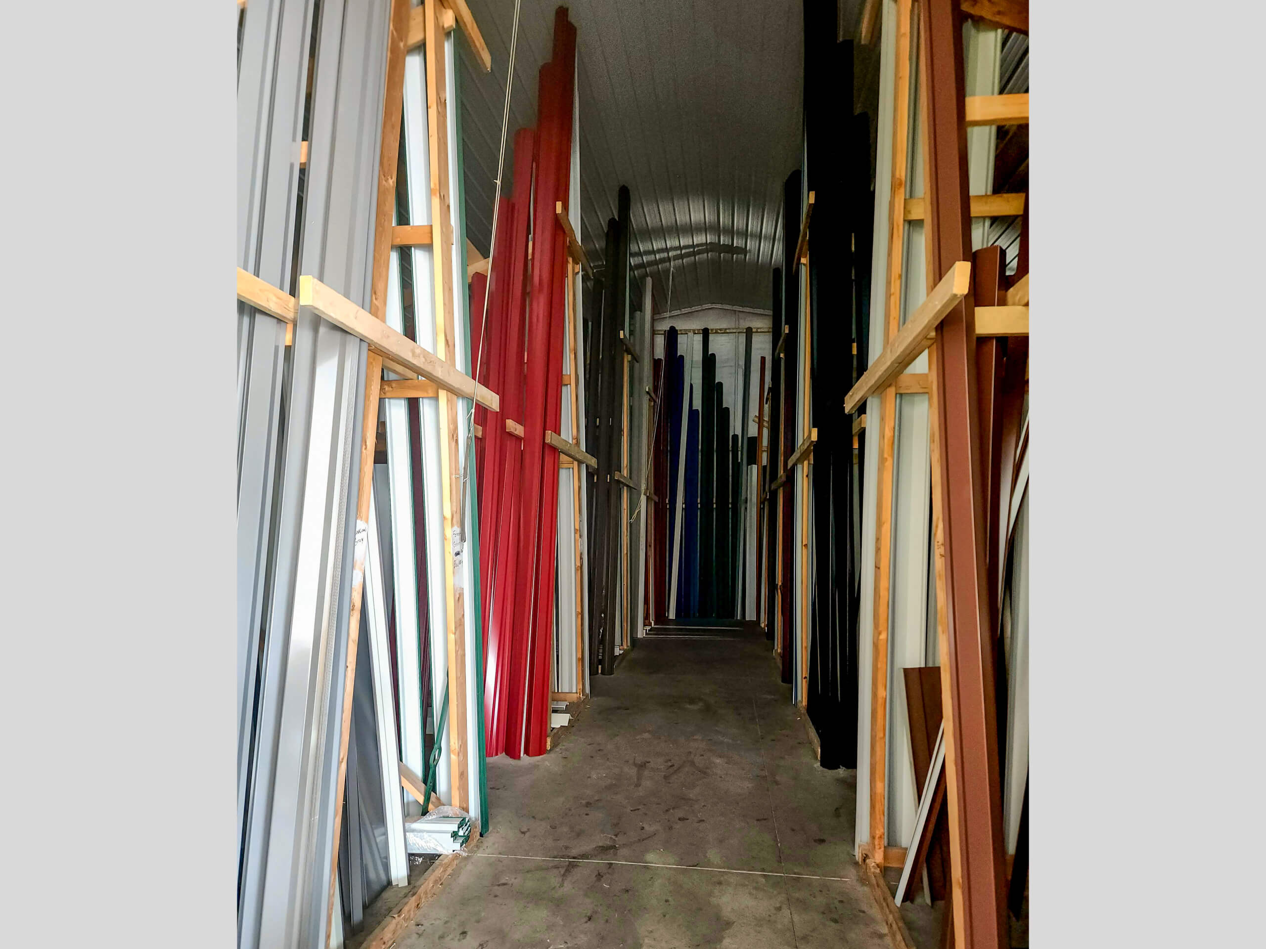 Sunset Metal Roofing and Siding Warehouse Ebensburg, PA Tall colored metal or plastic panels are stacked vertically on wooden racks along both sides of a narrow storage aisle with a concrete floor and arched ceiling.