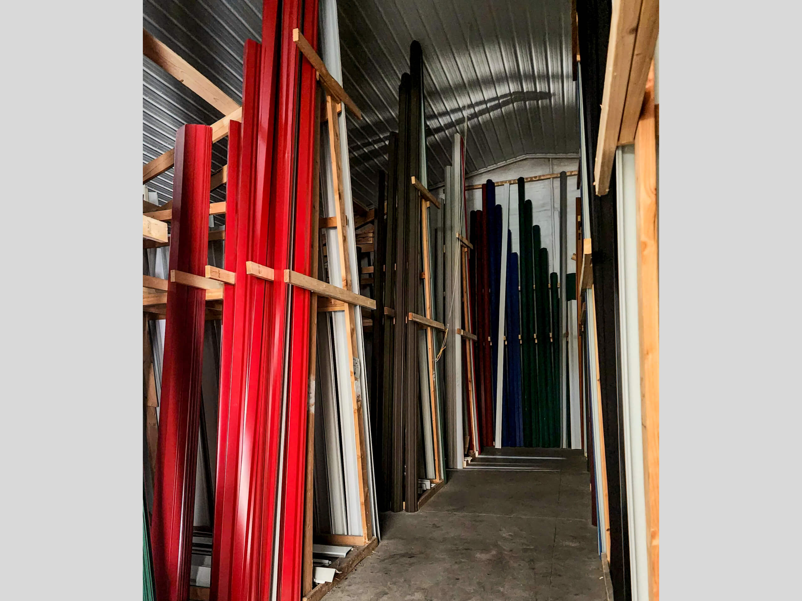 Sunset Metal Roofing and Siding Warehouse Ebensburg, PA Tall, narrow panels of various colors are stored vertically on wooden racks inside a metal-walled storage area with a concrete floor and a corrugated metal ceiling.