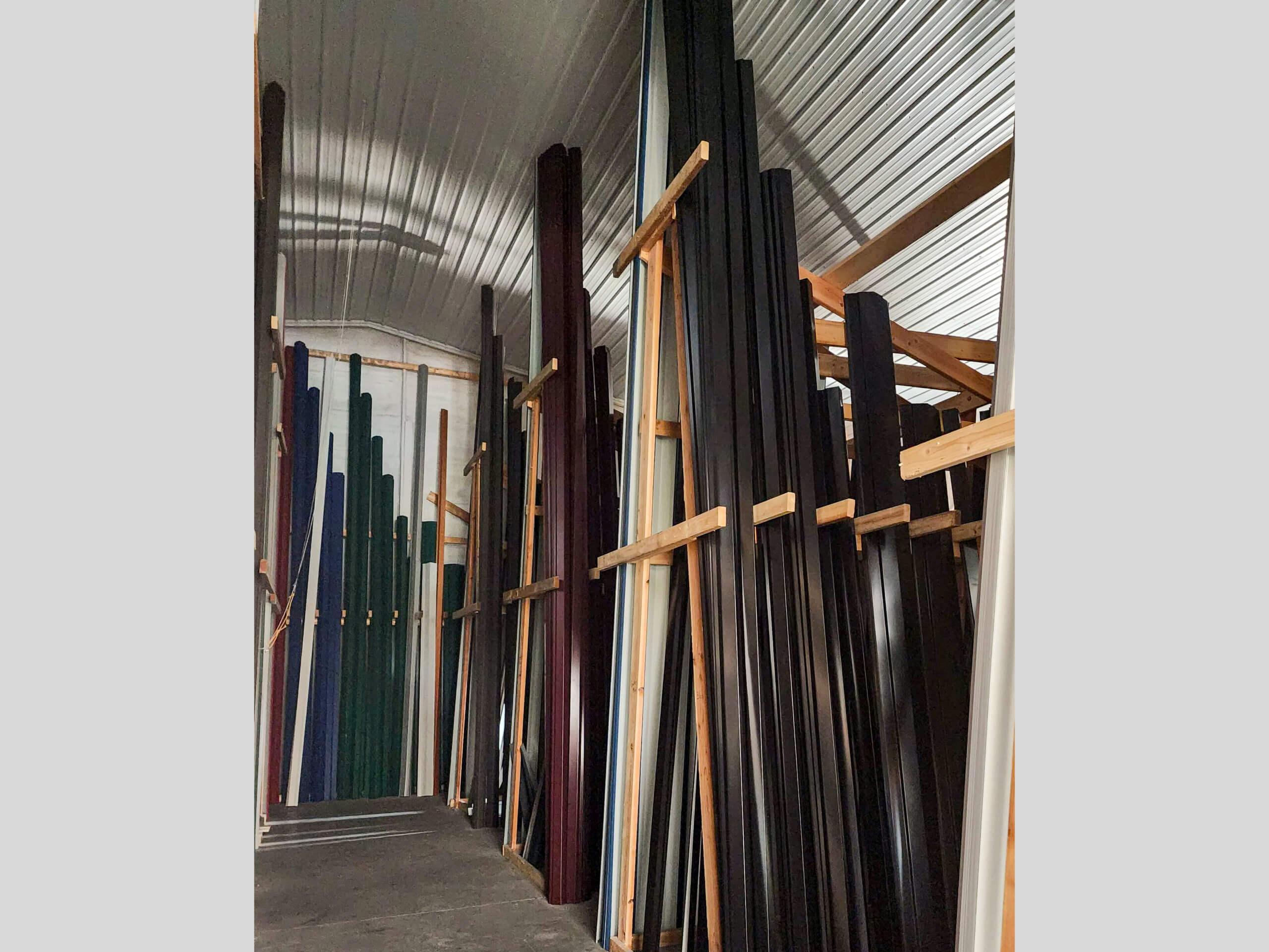Sunset Metal Roofing and Siding Warehouse Ebensburg, PA Tall metal or plastic panels in various colors are stored vertically on wooden racks inside a warehouse with a curved metal roof.
