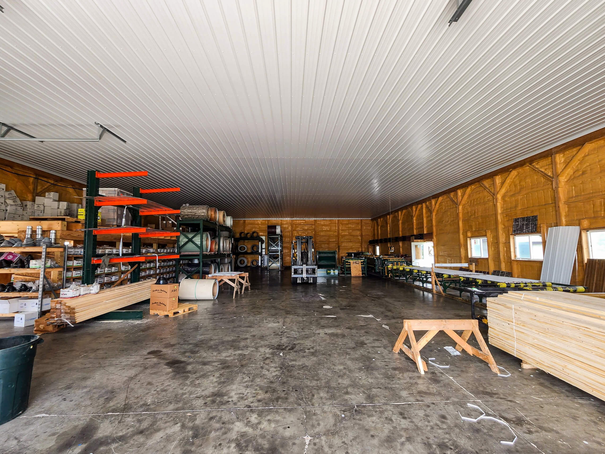 Sunset Metal Roofing and Siding Warehouse Ebensburg, PA Interior of a warehouse with stacked lumber, shelves holding supplies, and construction materials organized along the walls on a concrete floor.