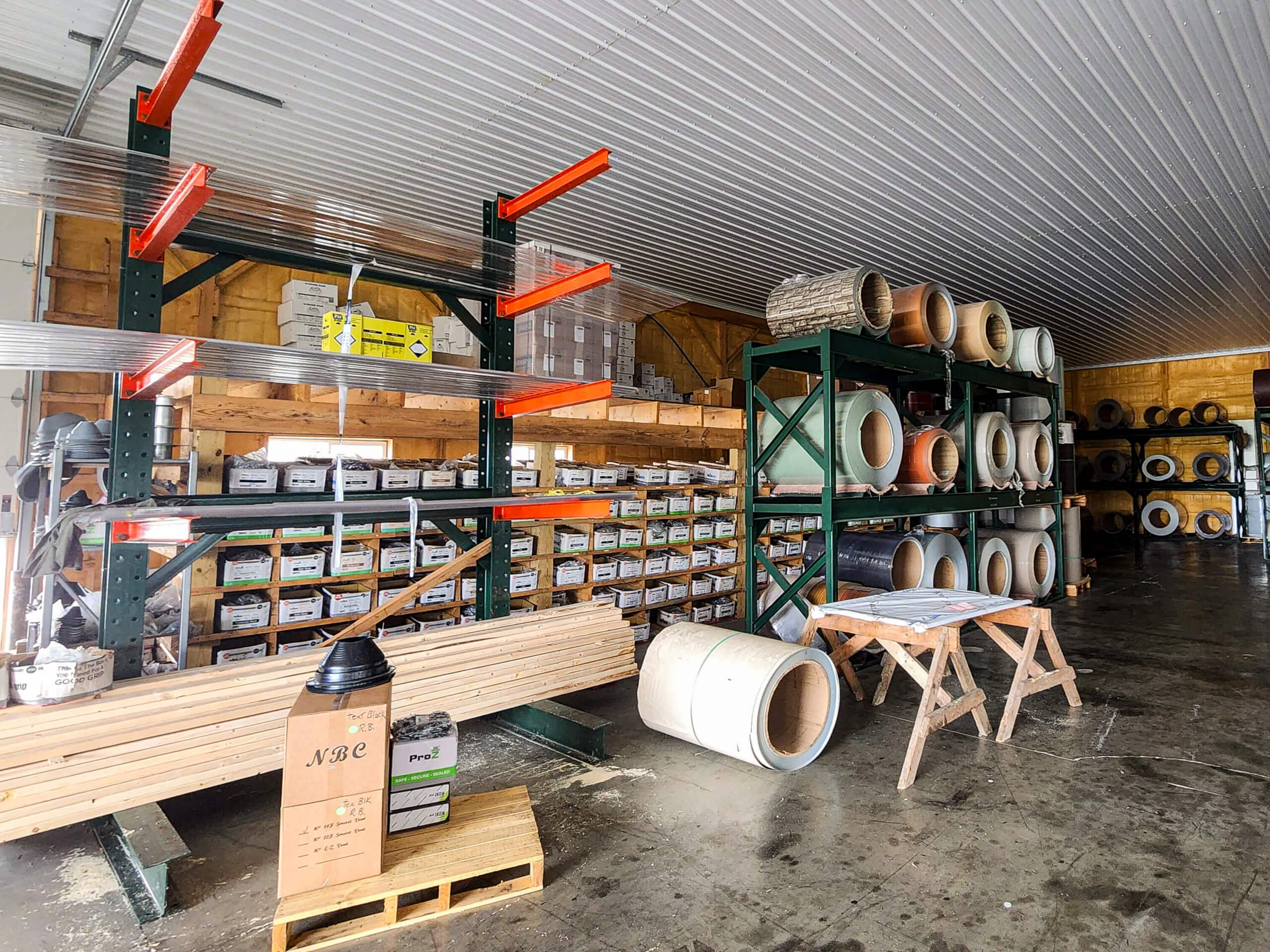Sunset Metal Roofing and Siding Warehouse Ebensburg, PA Warehouse interior with metal shelves holding boxes, spools of wire or cable, and stacks of wood planks on the floor. A saw horse and pallet with items are visible in the foreground.