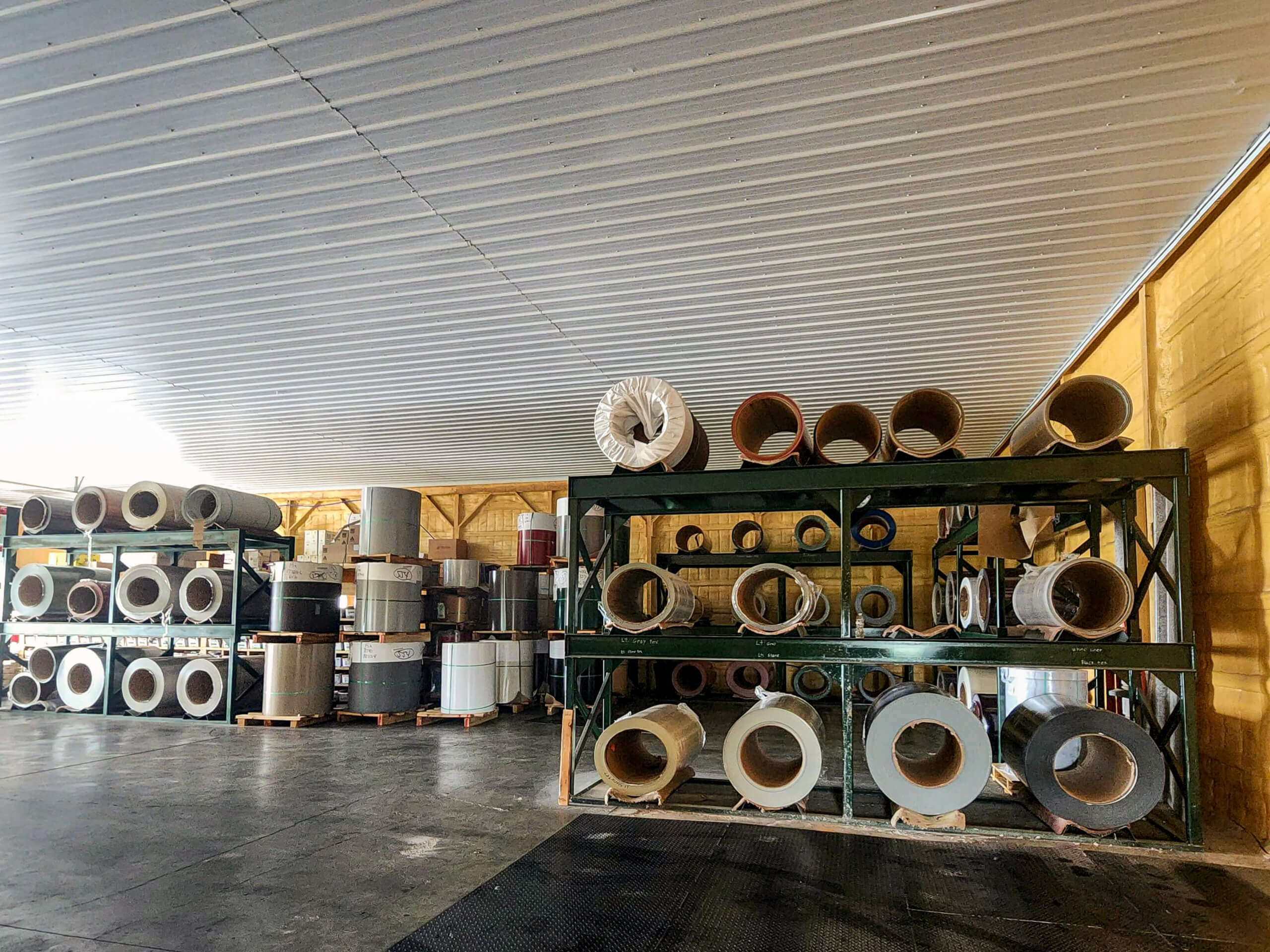 Sunset Metal Roofing and Siding Warehouse Ebensburg, PA Large rolls of industrial material are stacked on metal racks and wooden pallets inside a spacious warehouse with a corrugated ceiling.