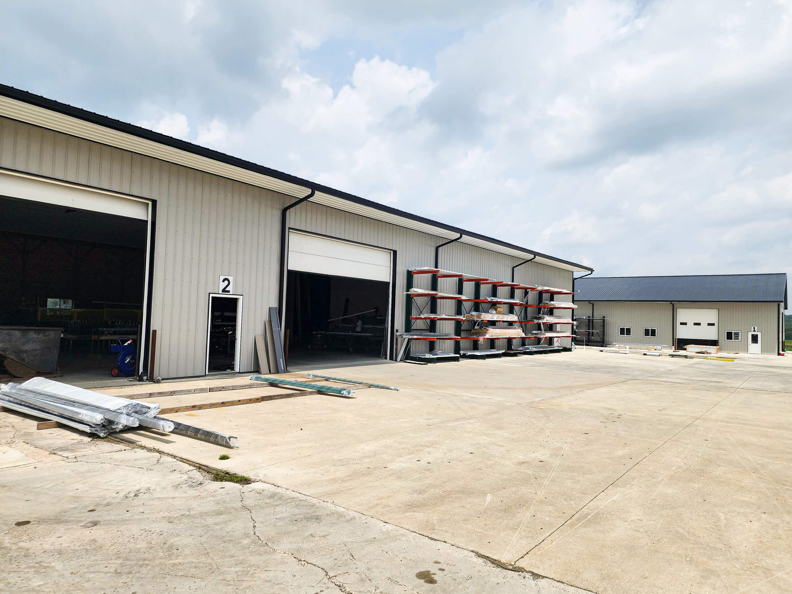 Sunset Metal Roofing and Siding Warehouse Ebensburg, PA Industrial warehouse with open garage doors, stacked pipes on racks, and metal materials on the concrete lot under a partly cloudy sky.
