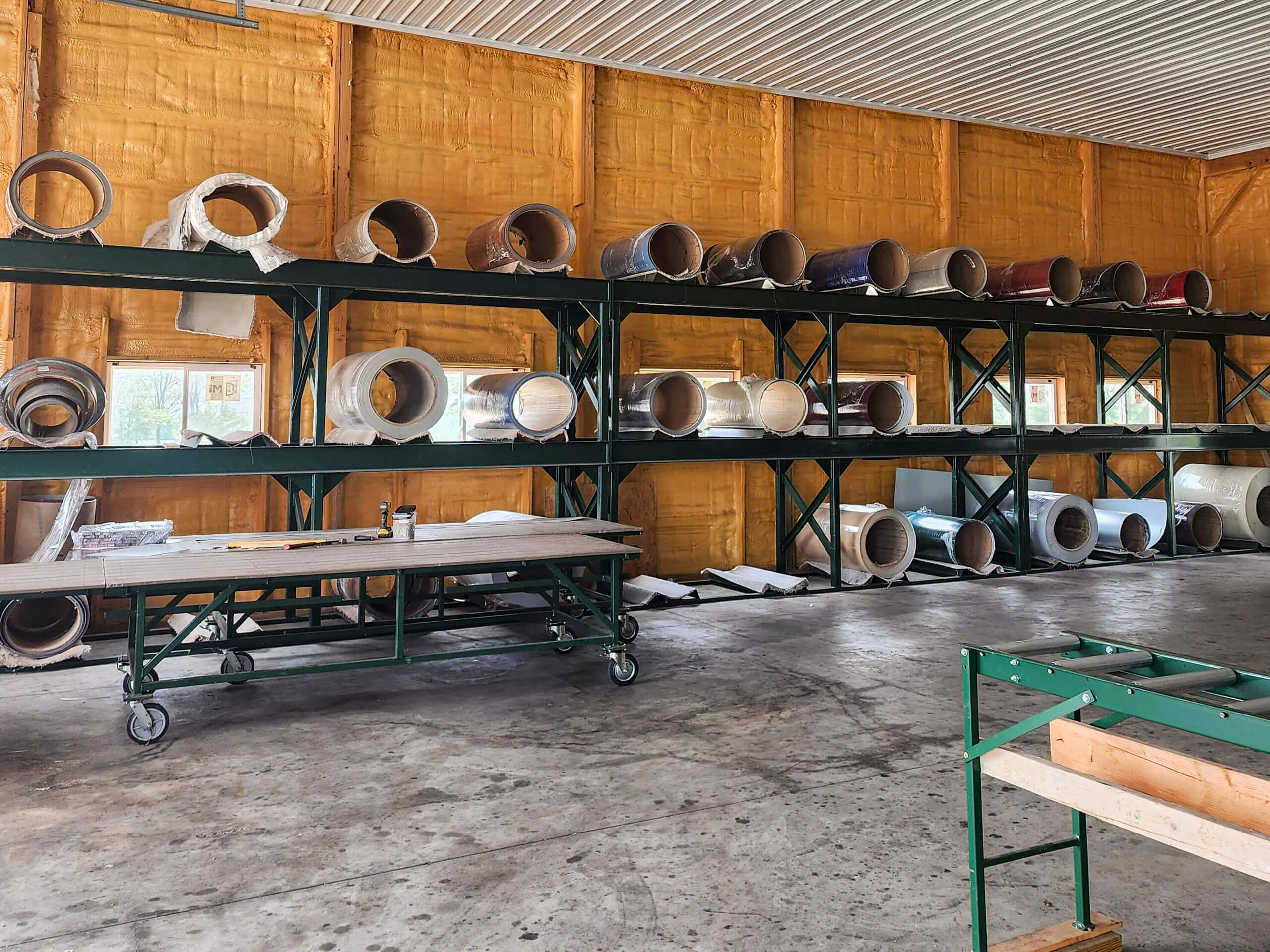 Sunset Metal Roofing and Siding Warehouse Ebensburg, PA Rolls of material are stored on metal shelves against a yellow insulated wall in a workshop. A worktable and green equipment are visible on the concrete floor.