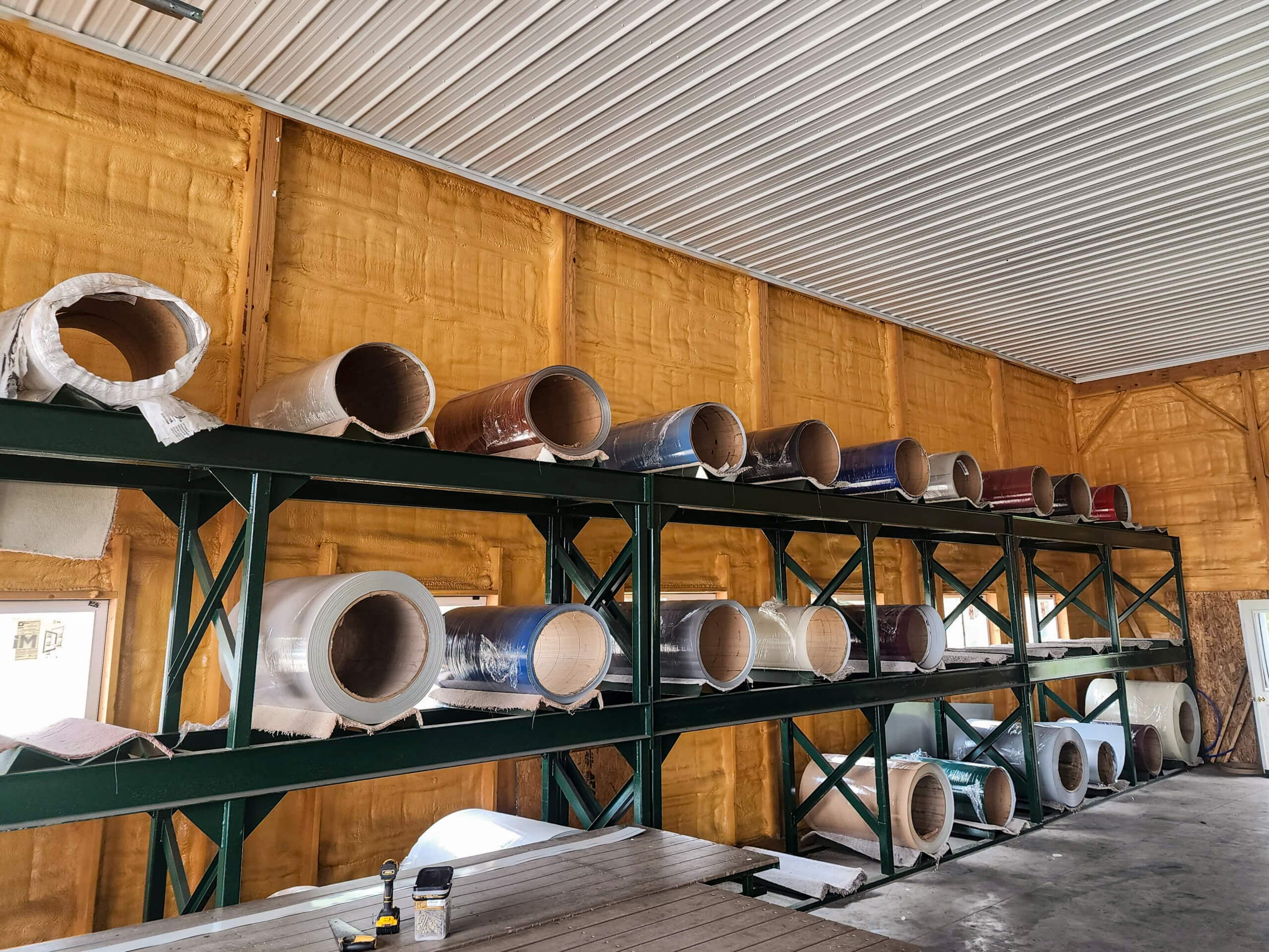 Sunset Metal Roofing and Siding Warehouse Ebensburg, PA Large industrial rolls of material are stored on green metal shelves against a yellow insulated wall inside a warehouse.