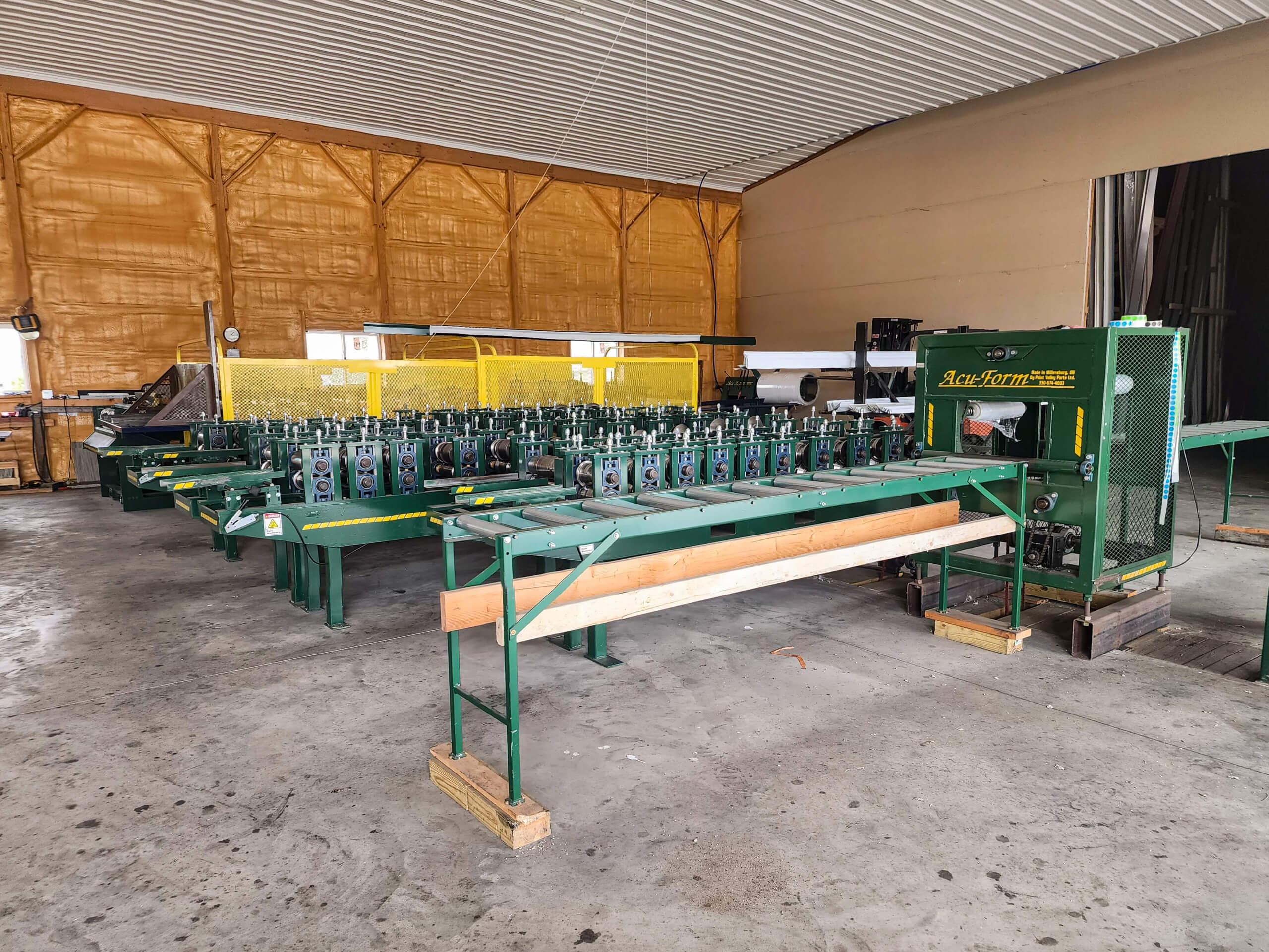 Sunset Metal Roofing and Siding Warehouse Ebensburg, PA Green industrial machinery, including conveyor belts and processing units, is arranged inside a spacious warehouse with yellow insulated walls and a concrete floor.