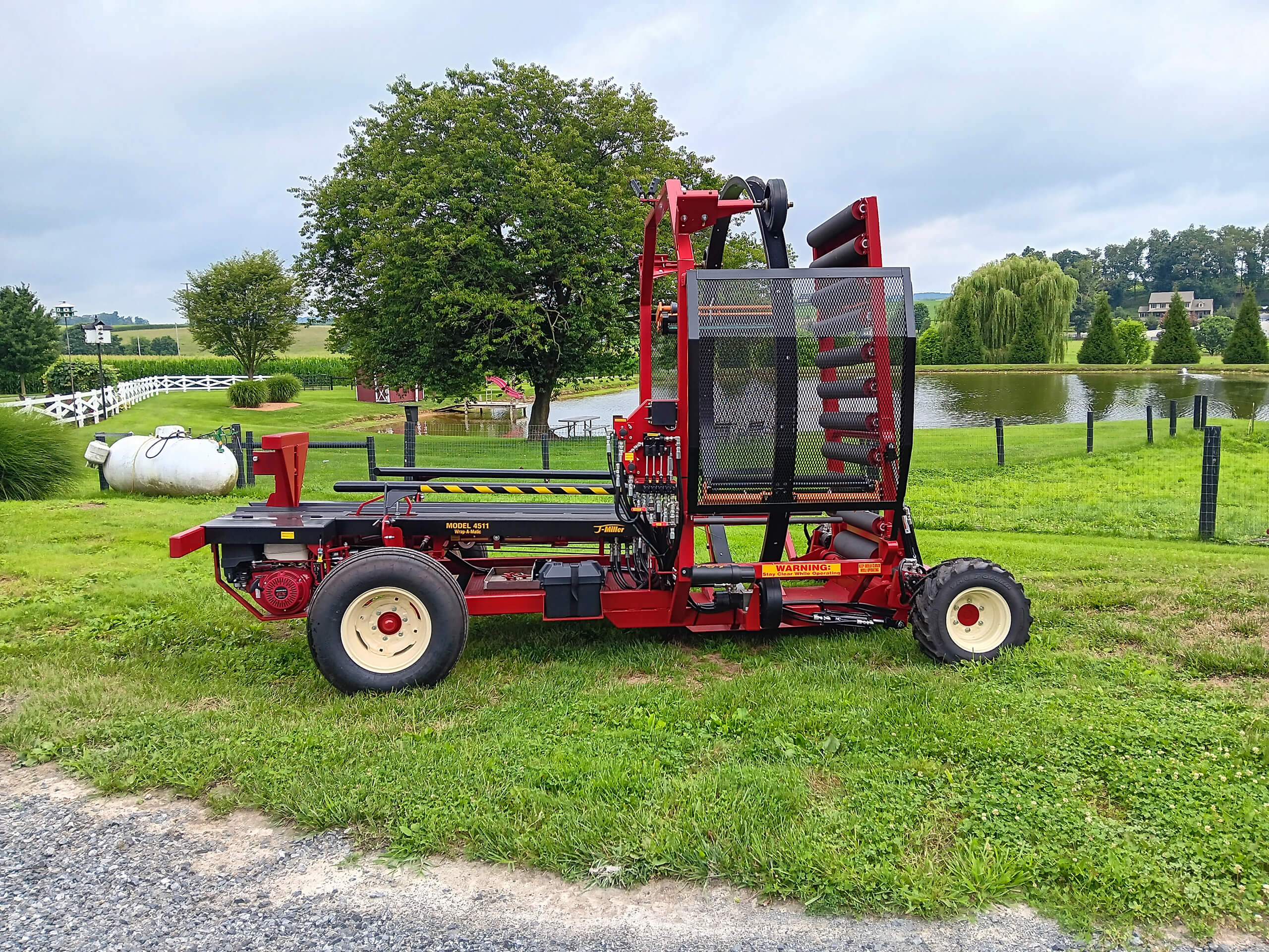 A red and black farm machine with white wheels sits on grass near a pond, trees, and fences under a cloudy sky.