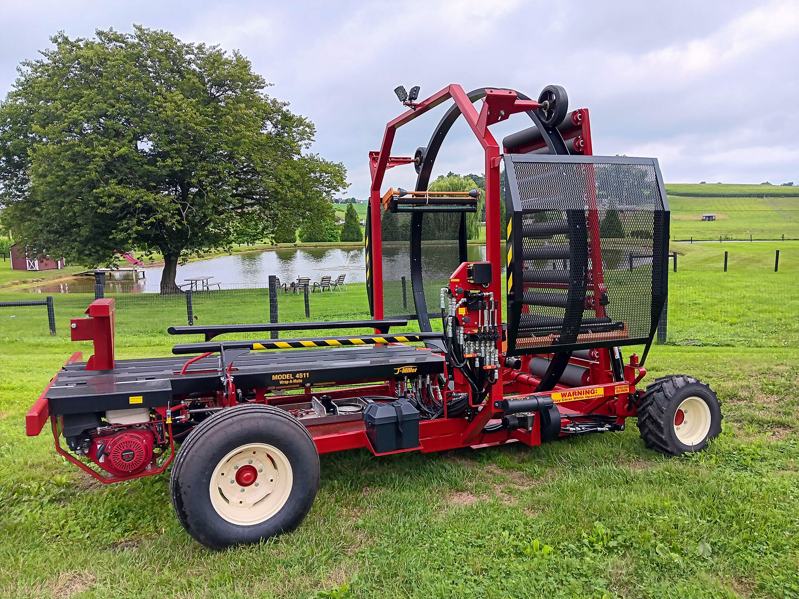 Red agricultural bale wrapper machine parked on grass with a pond, trees, and fence in the background under a cloudy sky.