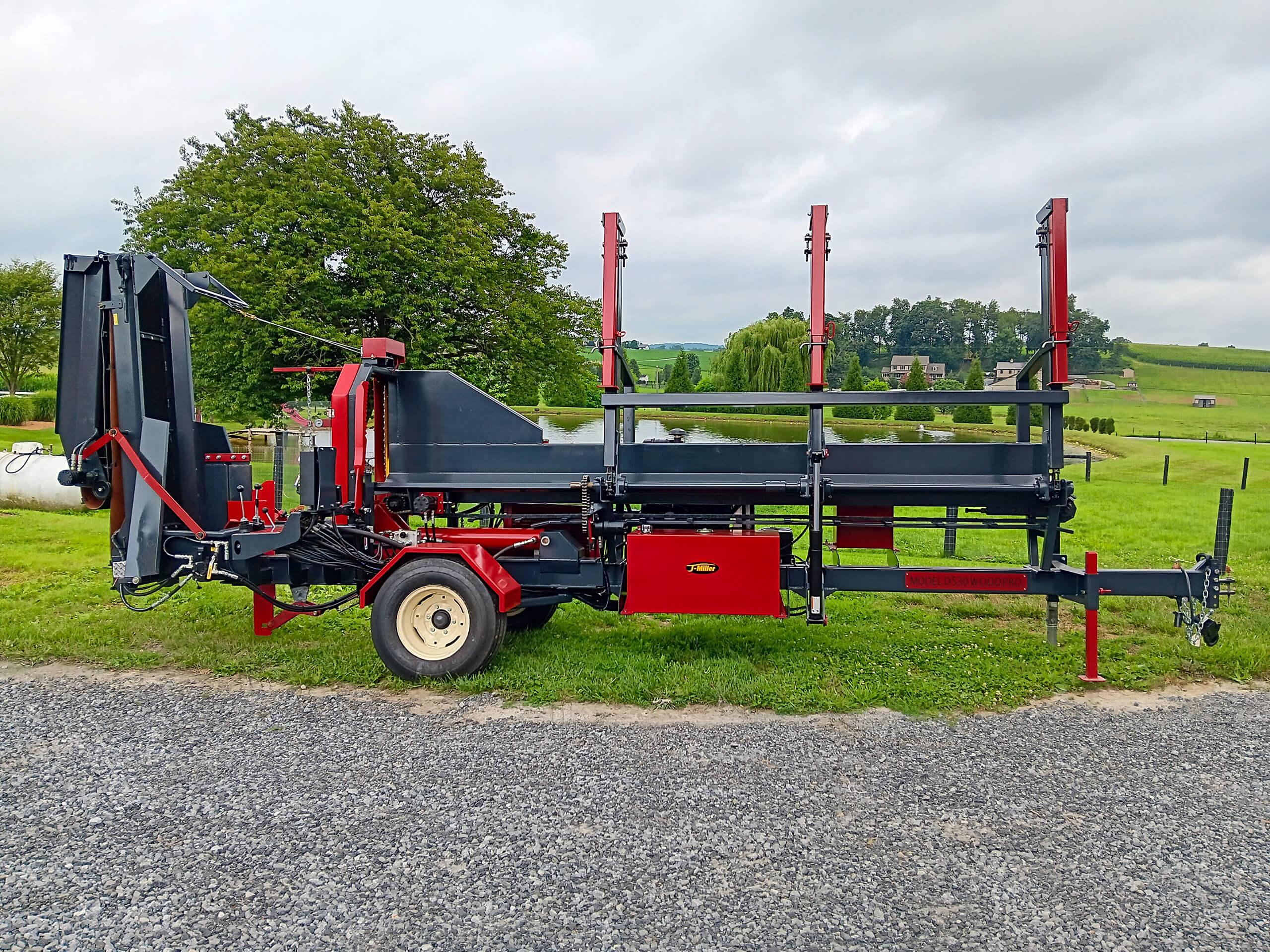 Red and black log processing machine with wheels, parked on gravel near a grassy field, trees, and a pond under a cloudy sky.