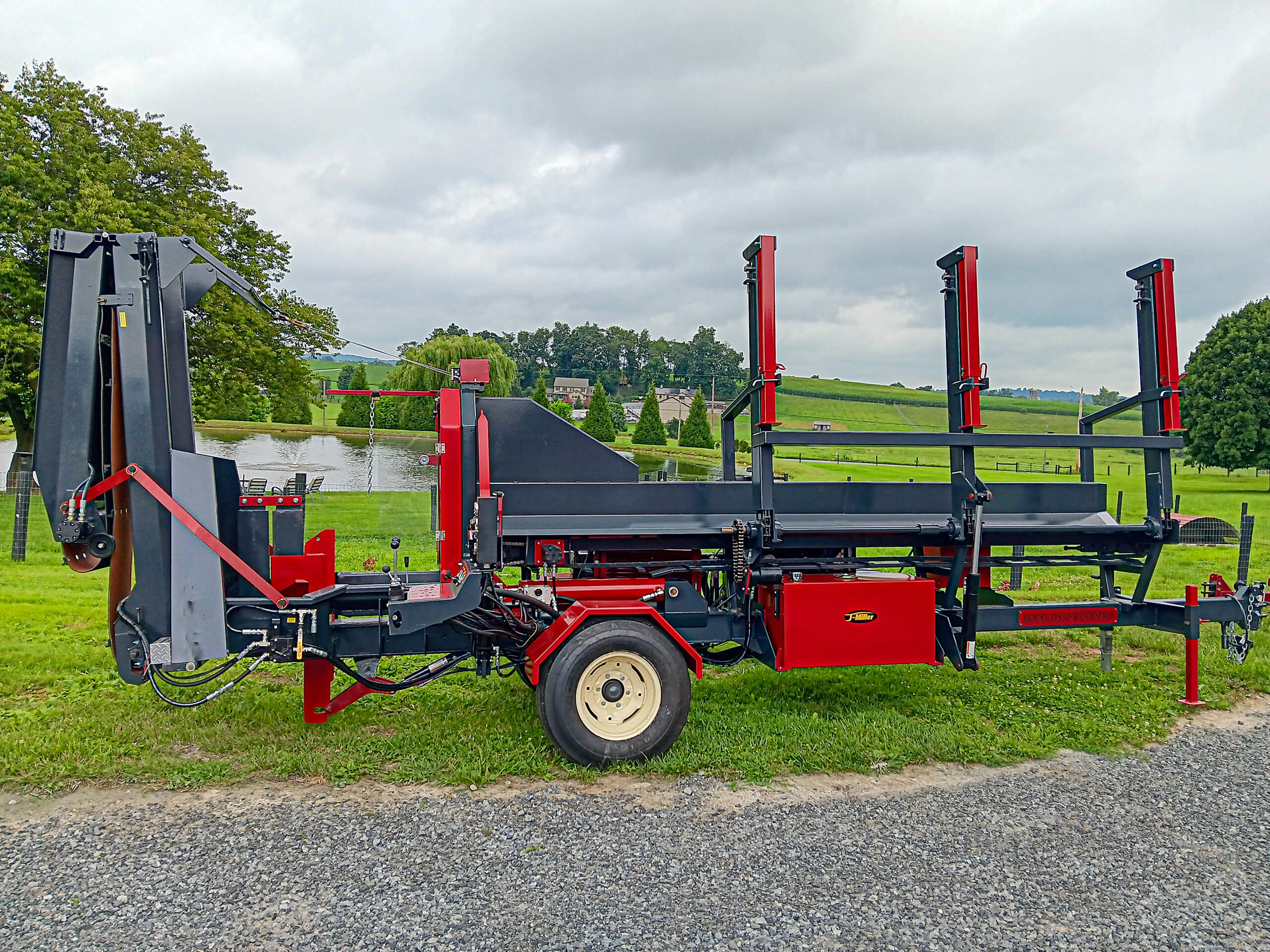 A red and black log splitter machine with vertical supports is parked on gravel, with green grass and trees in the background.