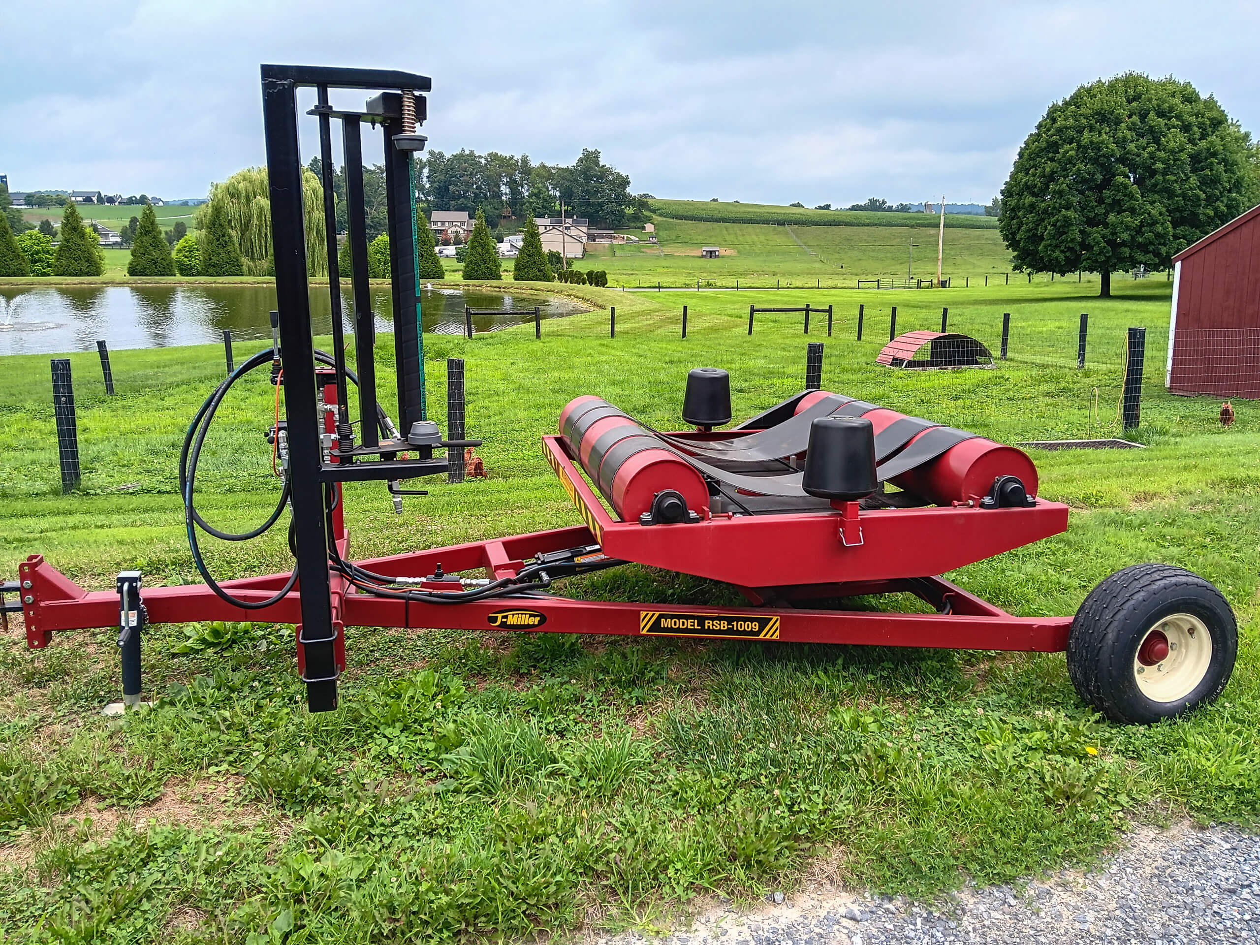 Red agricultural machine with rollers and hydraulic components, labeled "Model 1856-1009," situated on grass near a pond, fencing, and farm buildings in a rural landscape.