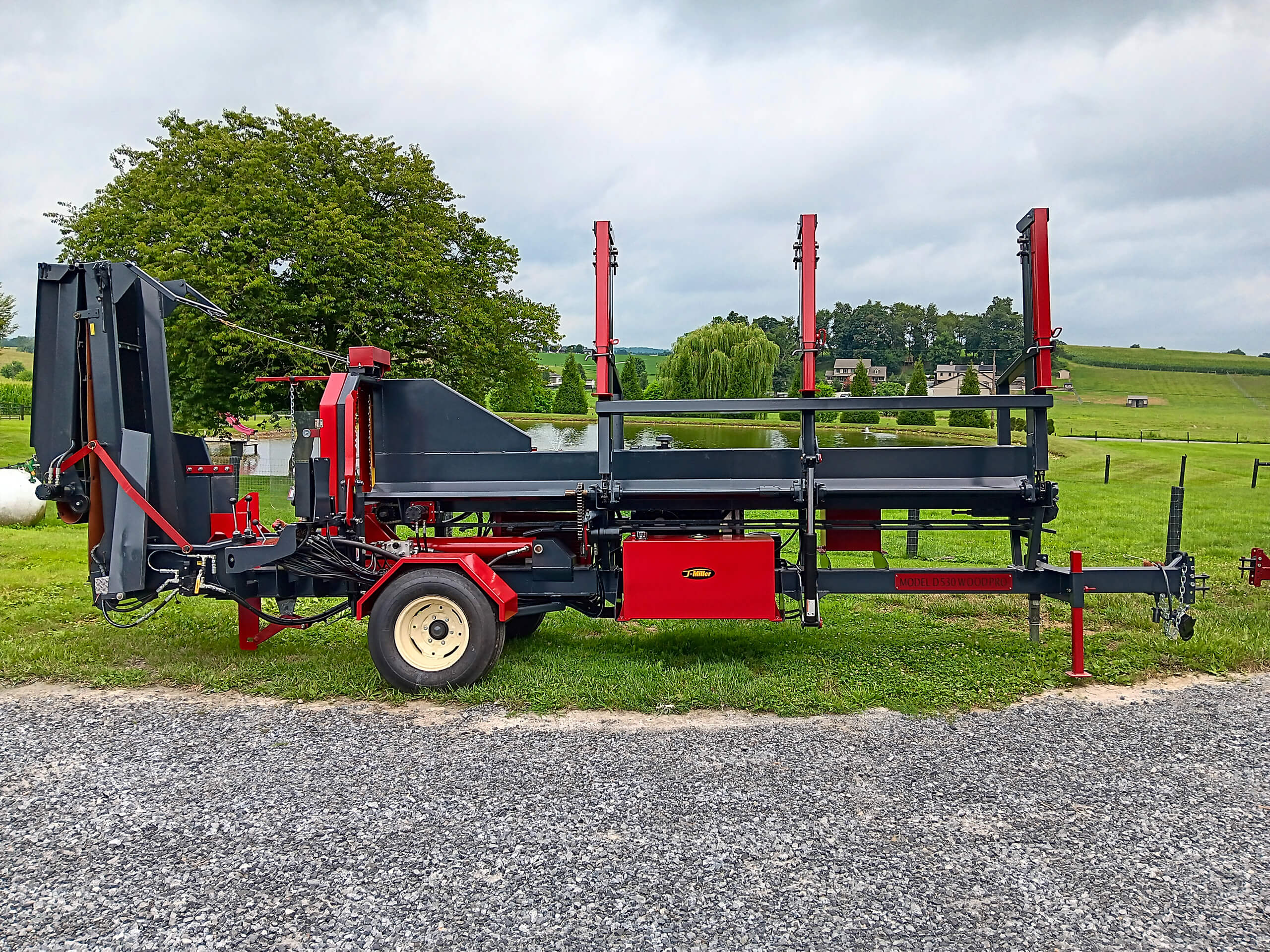 Red and black portable sawmill on gravel with grassy field and trees in the background under a cloudy sky.
