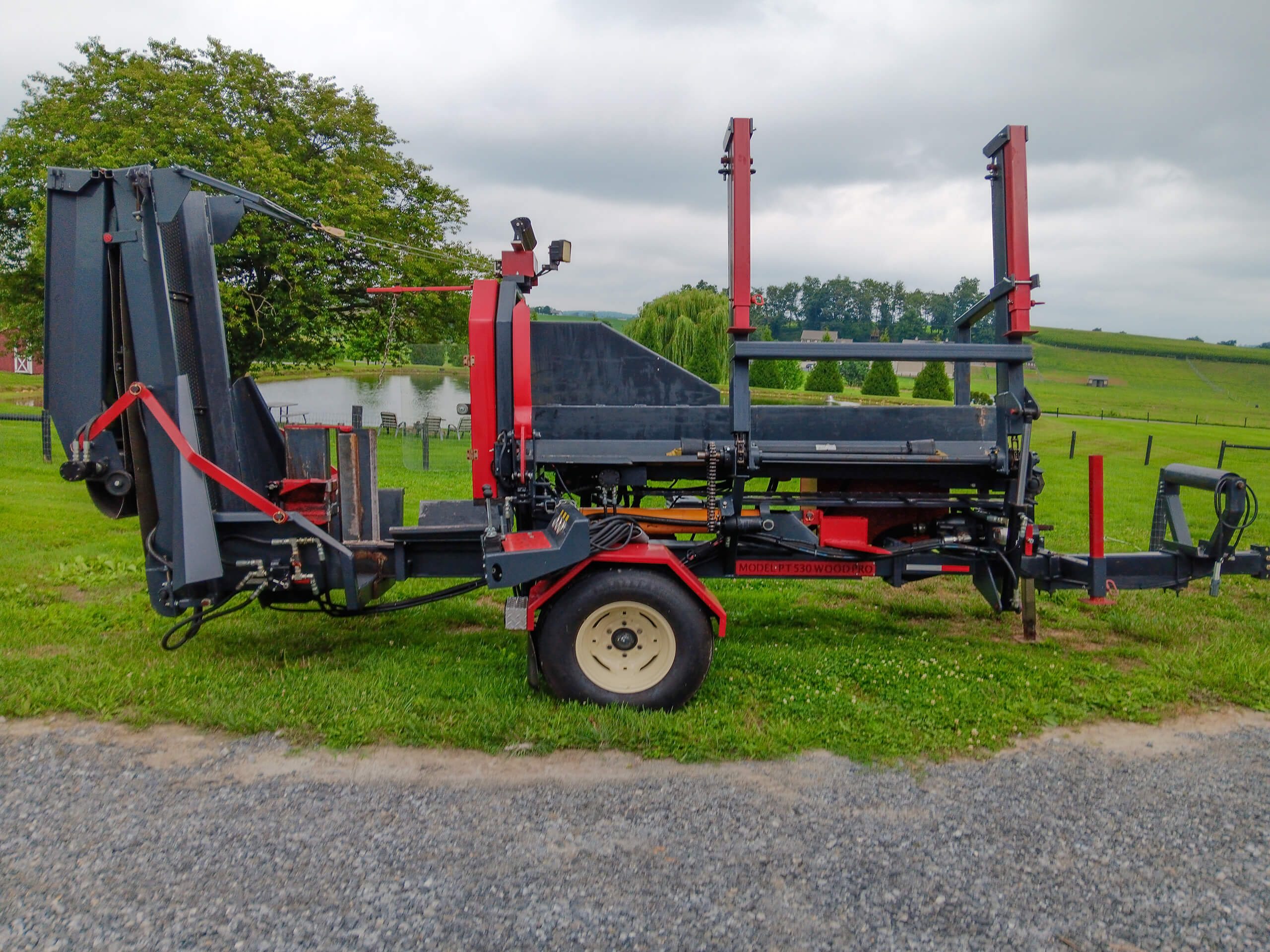 Red and black agricultural machinery on a grassy area, with a large wheel in the center and upright supports, set against a rural landscape with trees and a pond.